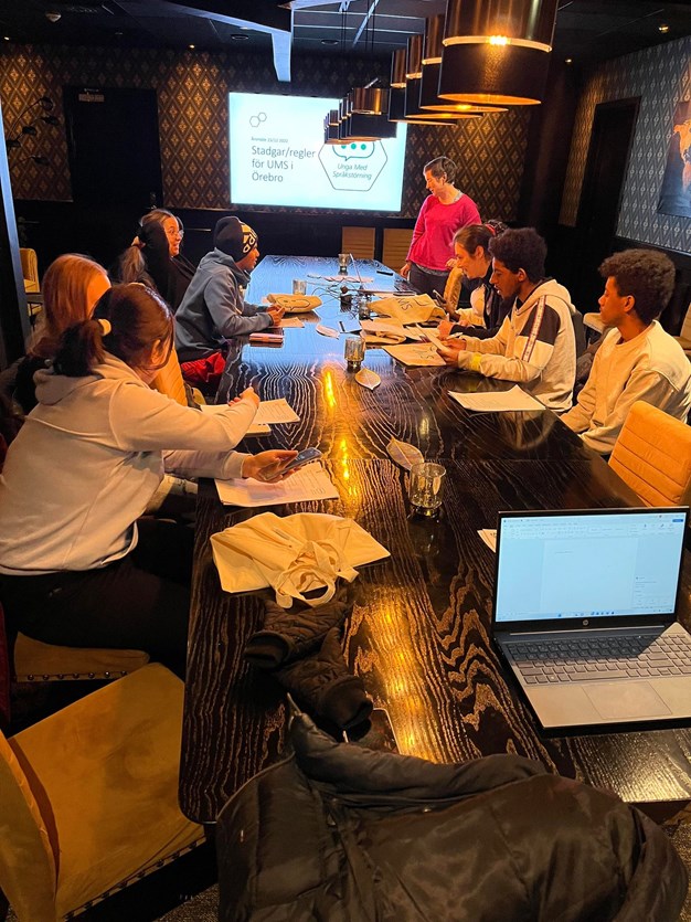 Nio personer runt ett mötesbord. en skärm som visar en presentation i bakgrunden